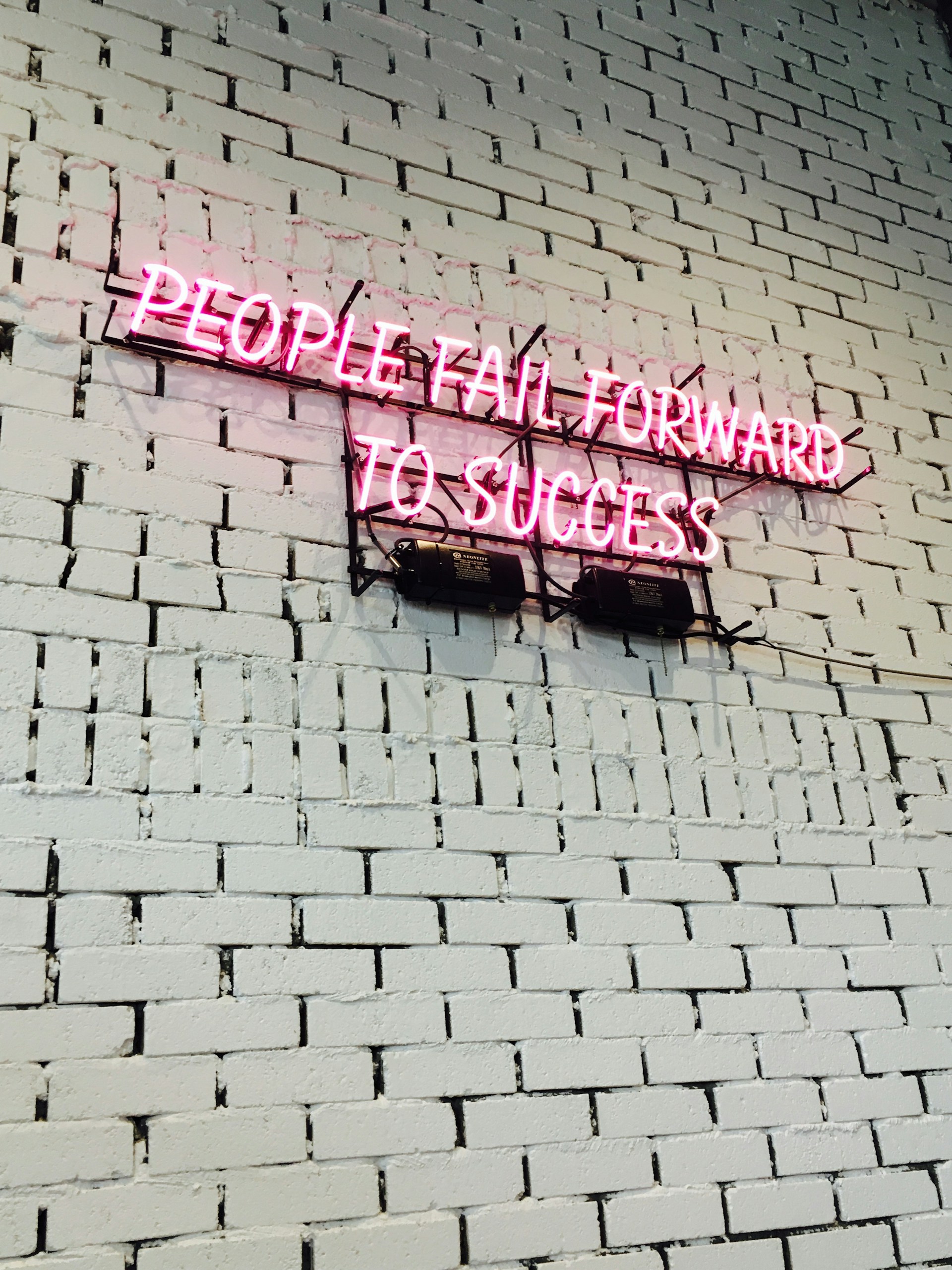 A pink neon sign on a painted brick wall that reads "people fail forward to success."