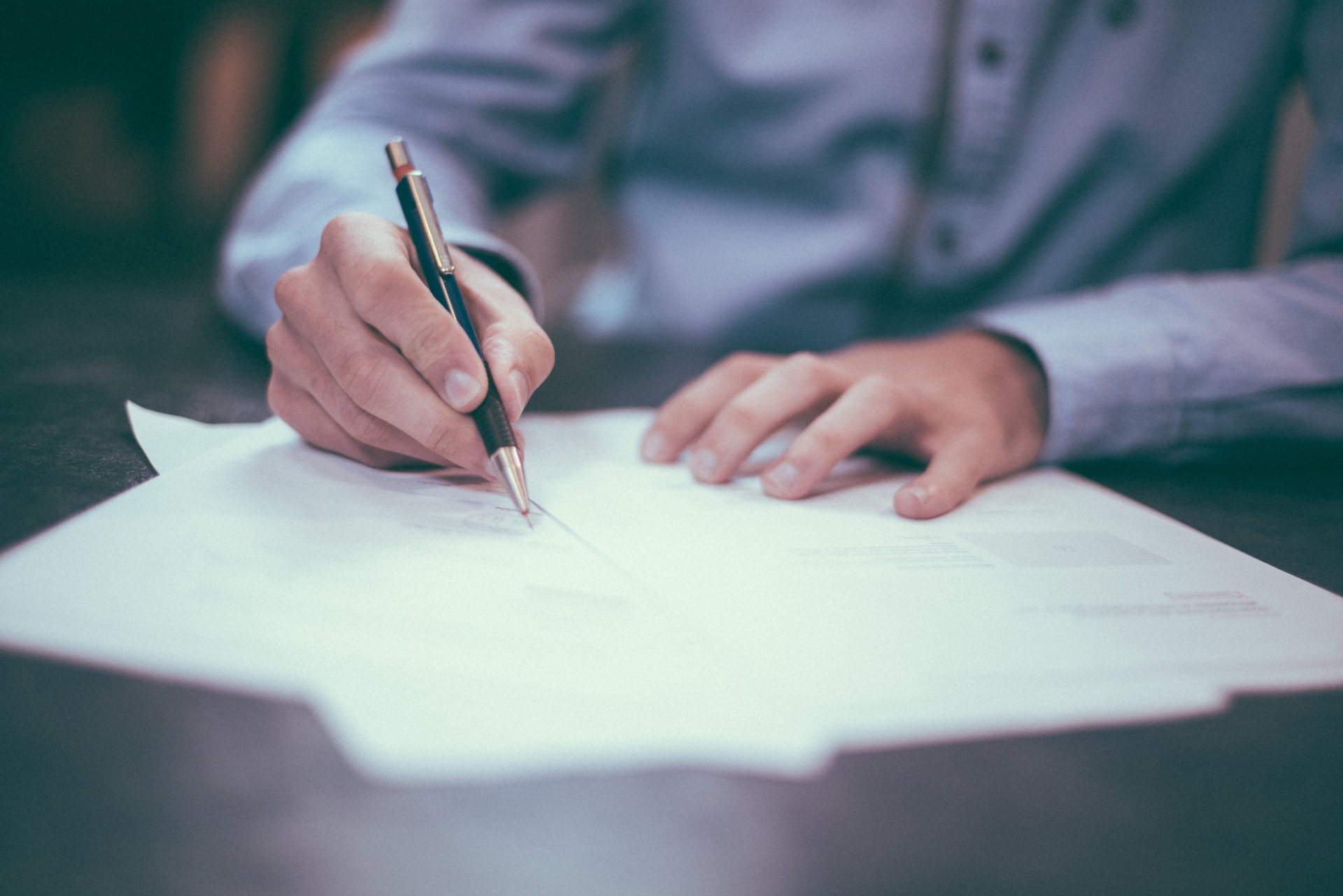 A person in business attire writing on several pieces of paper with a ballpoint pen.