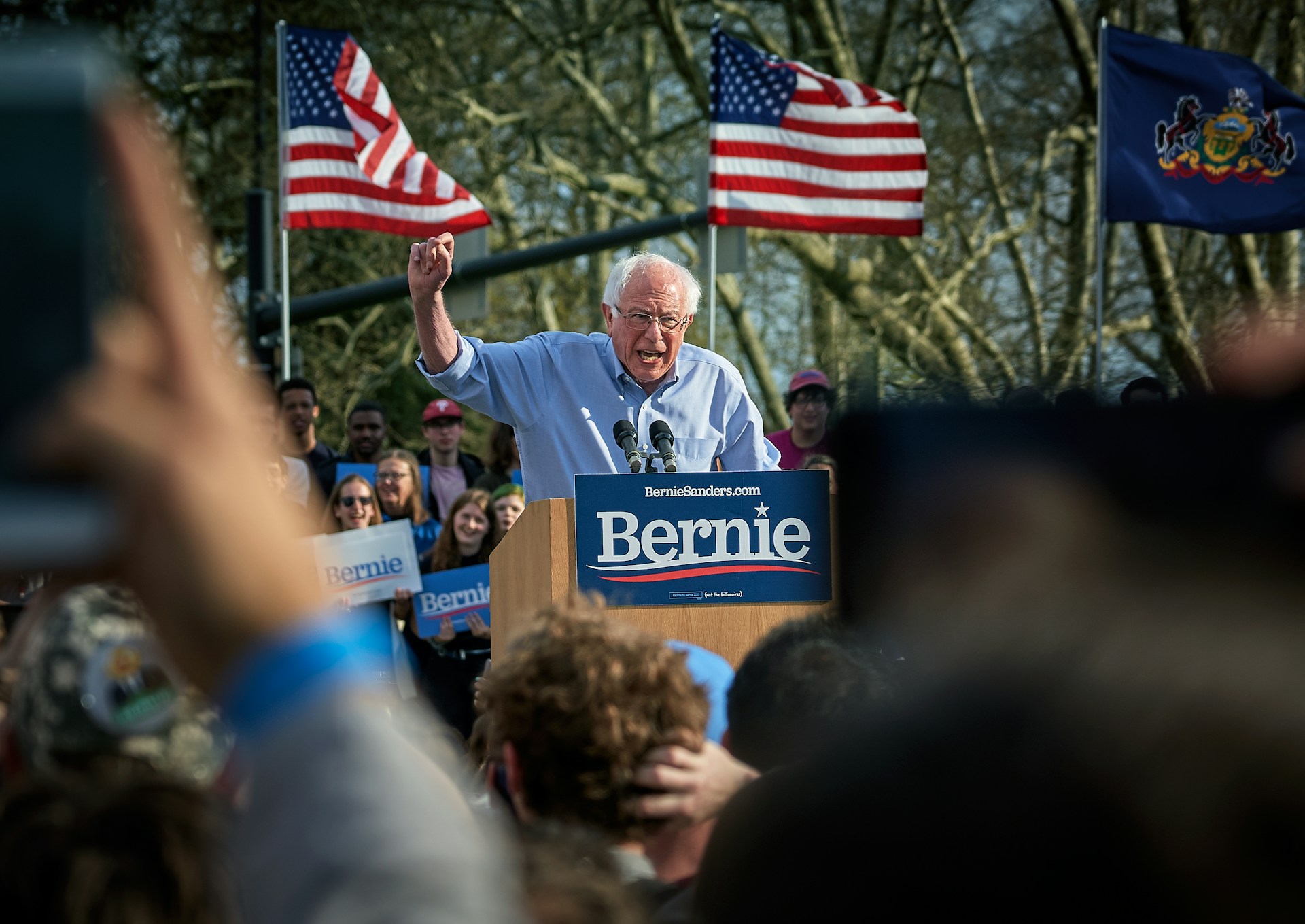 Politician Bernie Sanders speaking to a crowd from a podium set in the outdoors.